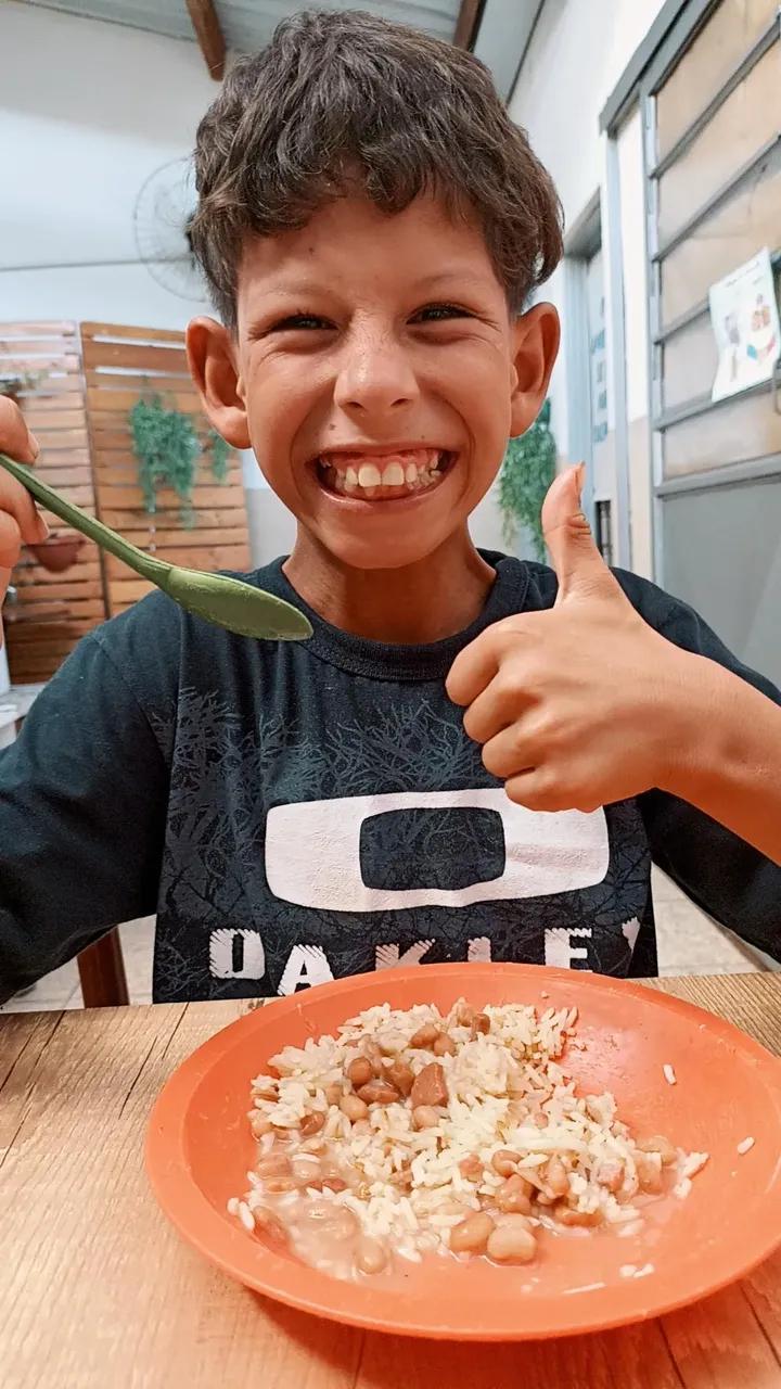 Menino sorrindo e fazendo joinha no refeitório, com prato de arroz e feijão